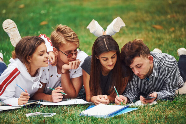 Writing on paper. front view. Group of young students in casual clothes on green grass at daytime.